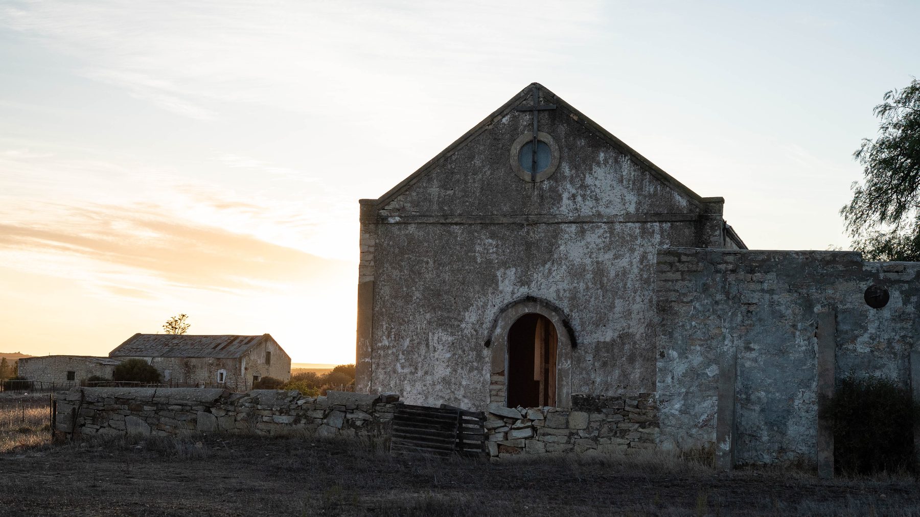 The original Nieuwoudtville church: a weathered stone building with a gable, round window and arched door, at dusk